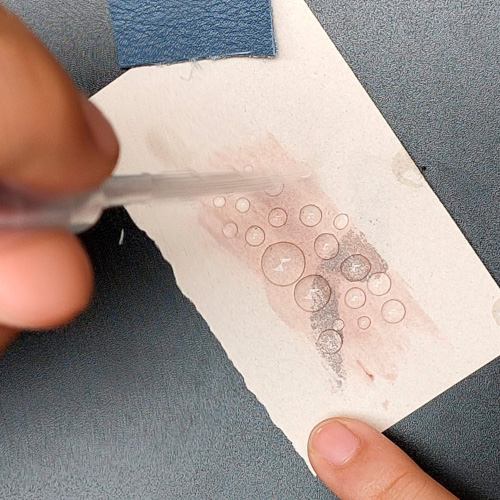 Vista detalle. Sobre un trozo de papel, una pipeta dispensa muestras de un líquido.  